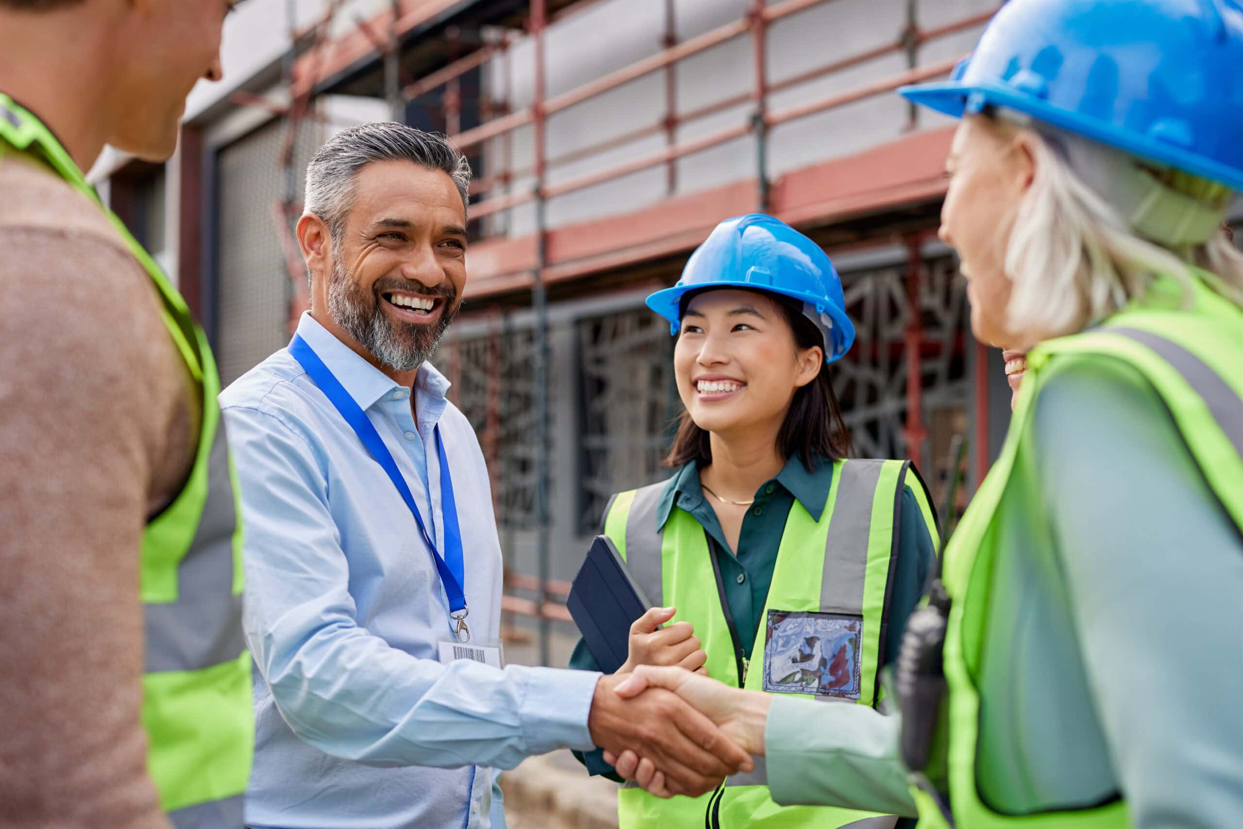 Zwei Bauarbeiterinnen und ein Bauleiter bei einem Baustellenmeeting, die sich die Hand geben, während sie auf einer Baustelle mit Gerüsten im Hintergrund stehen.
