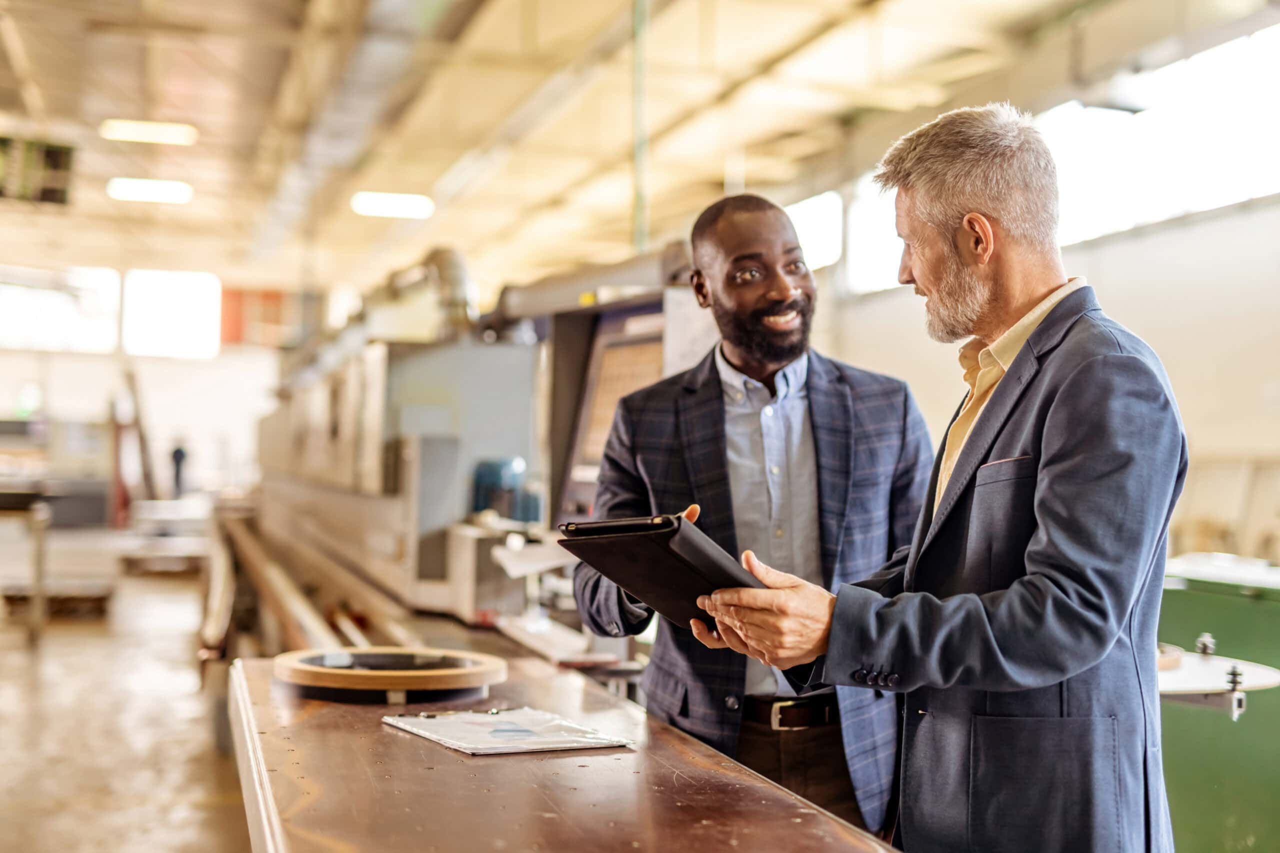 Dos hombres conversando en el suelo de una fábrica, uno de ellos señalando una tableta, símbolo de la contratación y el trabajo temporal en el sector industrial.