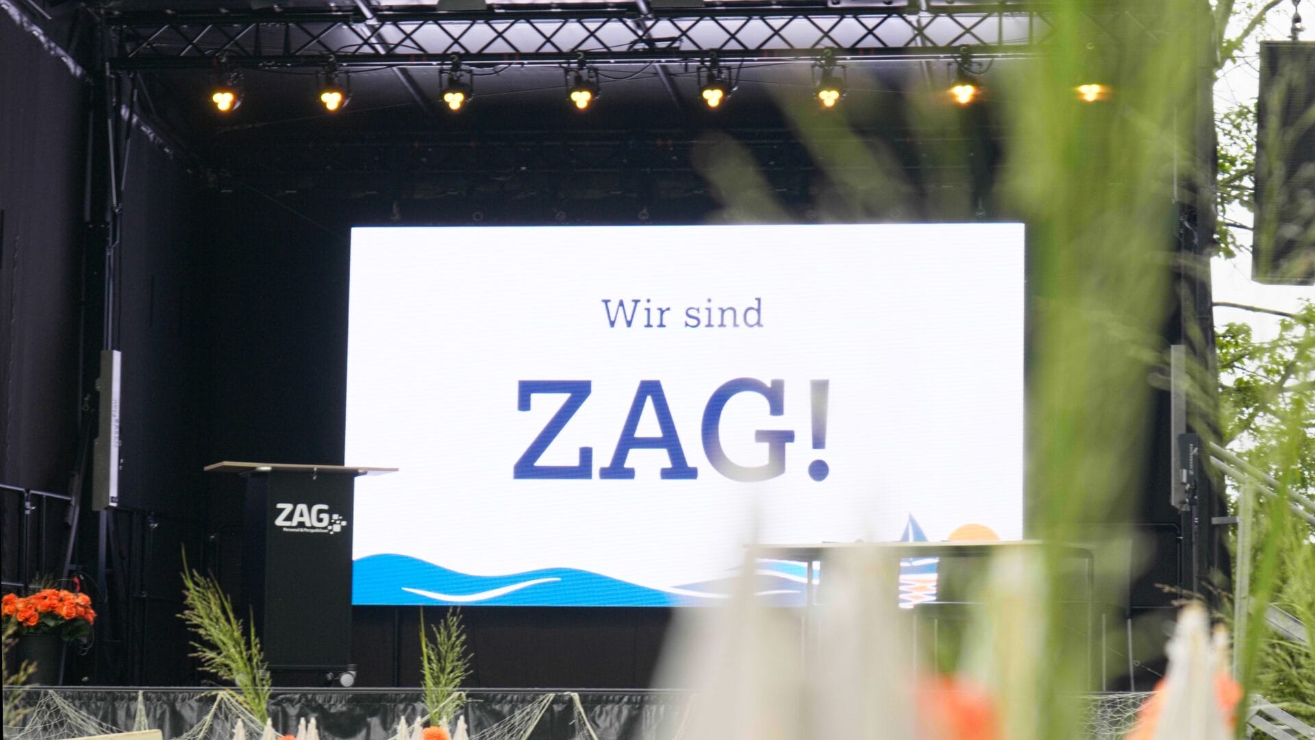 Grande logo ZAG su un palco in occasione della festa estiva del 2026, con lo slogan "Noi siamo ZAG!" sullo sfondo.