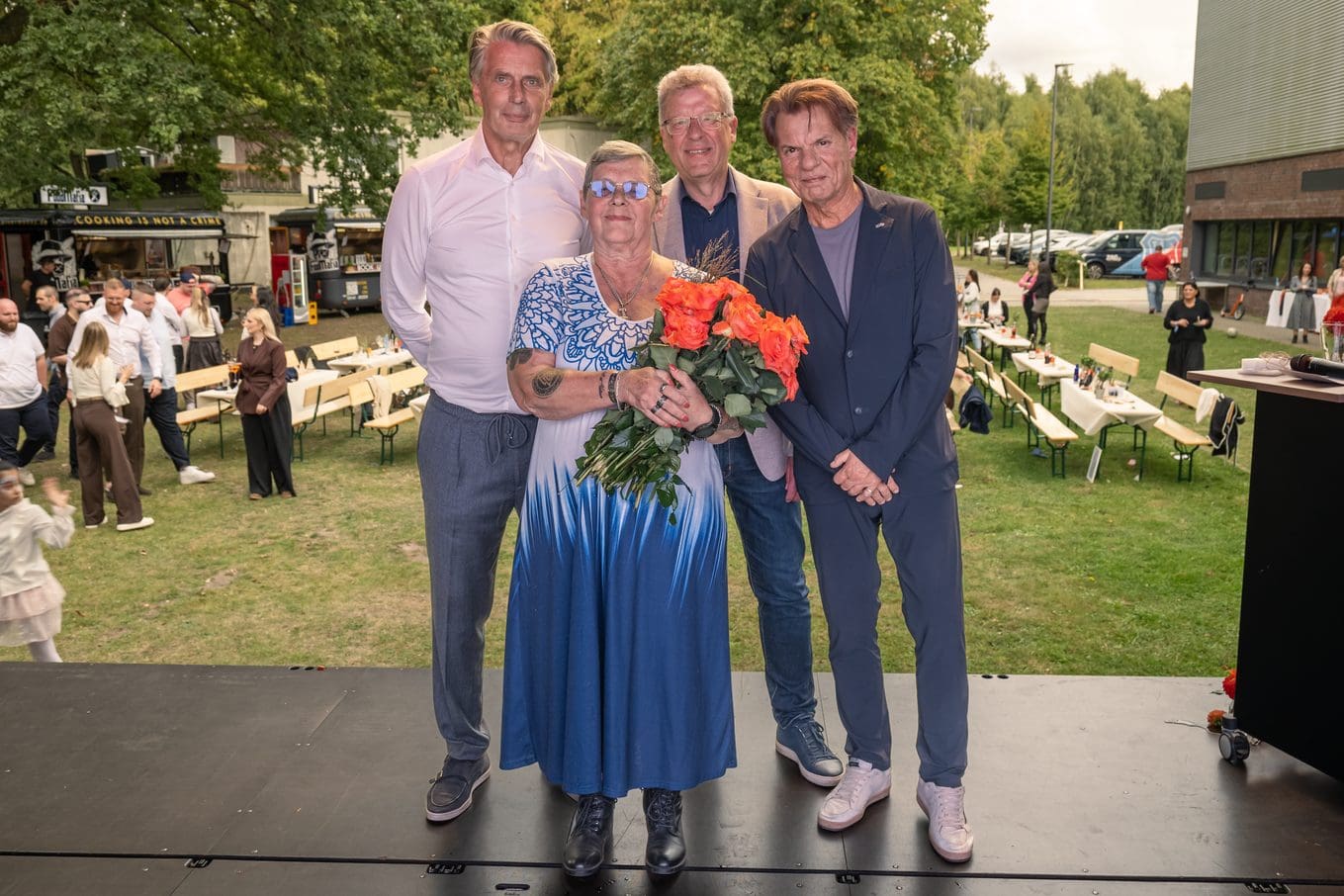 Quattro persone alla festa estiva dello ZAG del 2026, tra cui una donna con un mazzo di fiori, all’aperto con tavoli e ospiti sullo sfondo.