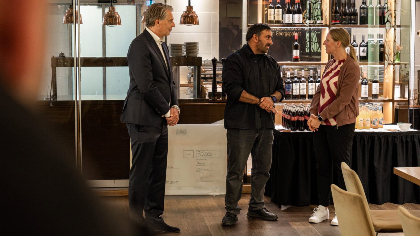 Zwei Männer und eine Frau beim Händeschütteln in einem modernen Veranstaltungsraum, im Hintergrund eine Bar mit Weinflaschen.